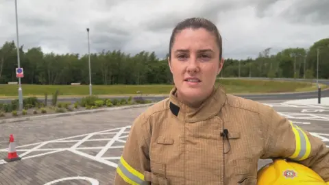 Caoimhe McNeice is young, with brown hair tied back and has brown eyes. She is wearing a tan coloured fire service uniform and has a yellow helmet under her arm.
