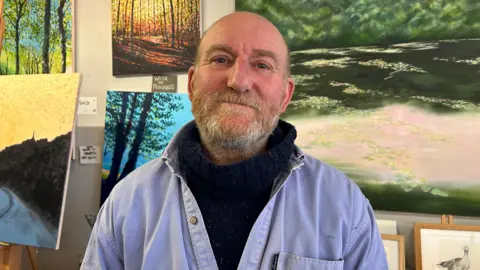 Man stands in front of paintings of trees and landscapes. He has a short beard. He's wearing a chunky black polo neck jumper and a blue overshirt. 