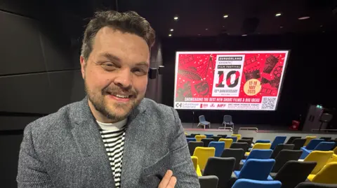 Chris Allan wearing a black and white striped shirt and a grey patterned blazer stands at the top of the auditorium, which has black, blue and yellow seating, while the cinema screen displays a red and white-coloured graphic showing the film festival is in its tenth year.