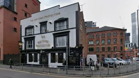 Google Image hows the Britons Protection, a three-storey pub rendered in white, with black outlining on the doors and window frames. It is surrounded by some Victorian era warehouses and some newer build, smaller apartment blocks