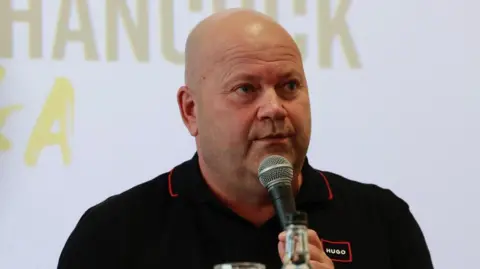 A man wearing a black t-shirt speaks into a microphone.