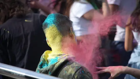 A splash of pink paint flies towards a boy. The back of his head is covered in yellow and blue. His black hoodie is also covered in blue and yellow.