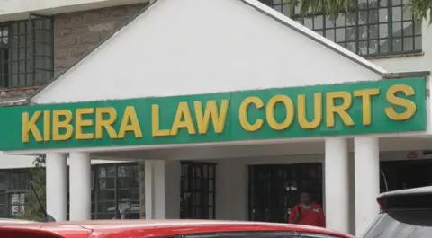 Two storey brick building with black window frames and large white pediment, with Kibera Law Courts written in yellow on a green background. 