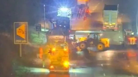 National Highways A blurred image of workers and heavy machinery working on a road. It is dark outside but there are lights on the vehicles. There are lorries and tractors on the road.