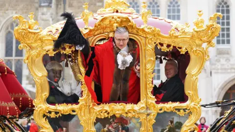 PA Media The Lord Mayor, dressed in ceremonial robes with a chain of office, leans out of a grand, gold gilded state carriage adorned with intricate decorations and paintings. He holds a black feathered hat and smiles at the crowd. Another official, also in traditional attire, sits beside him, while a another in a wig and white gloves waves from inside the carriage.