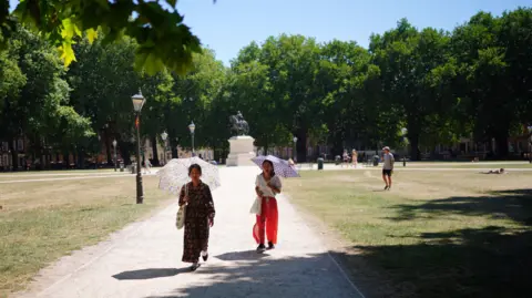 PA Media Two women hold umbrellas to shield themselves from the sun as they walk down a path in a park.
