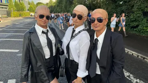 Three women all wearing white shirts, black ties, sunglasses and bald caps. The two on the outside are wearing black jackets.