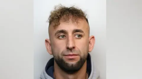 A mugshot of a man with a dark beard mainly around his chin. He is wearing a grey hoodie and looking to the side. He has frizzy dark blonde hair on the top of his head only. 