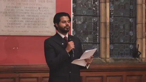Oxford Union/YouTube Moosa Harraj speaks to the Union holding a microphone and sheet of paper. He is bearded and wears a dark suit. Behind him is a stone plaque on the wall and a stained glass window.