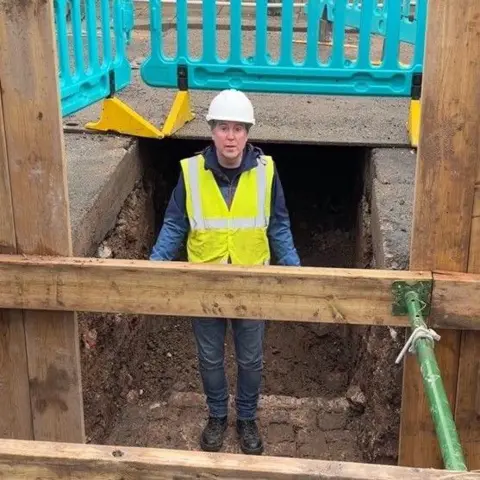 Hull City Council A man in jeans, black boots, a blue fleece, yellow hi-vis vest and white helmet is standing on bricks at the bottom of a trench, with his head at ground level. Wooden panels can be seen in front of him and blue barriers can be seen at road level.