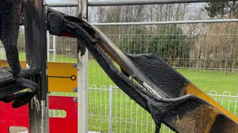 Billy McAloon A badly burned yellow slide, the metal of which has become warped and corroded. A fence has been put up around the slide.