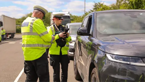 Two police officers holding a device next to a stalled car with two people inside