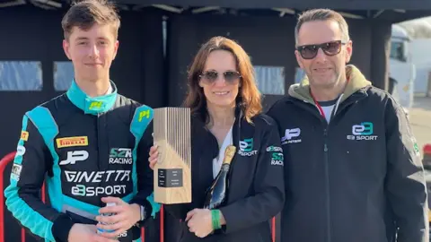 Kirsty Peters Jude in blue and black race suit, next to his mum, who is holding a bottle of champagne and a trophy, and his dad to her right. Both parents have a jacket promoting Jude's racing team.