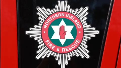 A crest of the Northern Ireland Fire and Rescue Service is shown on the side of a fire engine. The crest has a red hand in the centre surrounded by a white star and a green circle extending around it. The name of the organisation is encased further out around the crest.