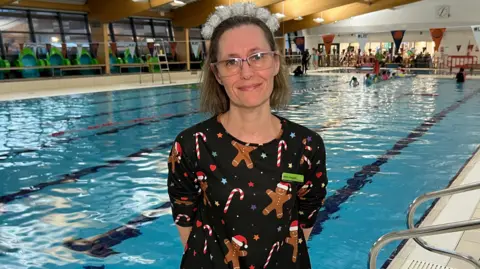 Jenny Hughes. She has glasses and short blonde hair and is pictured in front of a swimming pool. She is looking at the camera and smiling.
