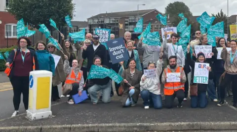 A group of dozens of people are at a road junction, with many holding light blue banners. Some are kneeling down and others are standing up, with buildings and trees in the background.