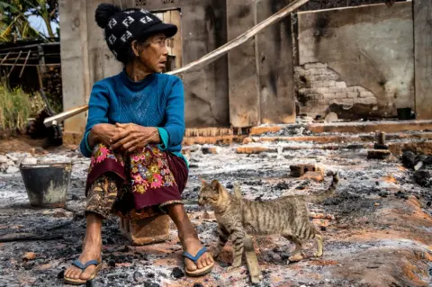 Getty Images This photo taken on February 4, 2024 shows a woman sitting next to a cat in front of her house damaged following fighting between Myanmar's Military and the Kachin Independence Army (KIA) in Nam Hpat Kar, Kutkai township in Myanmar's northern Shan State.