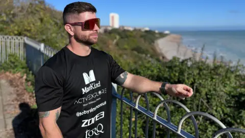 Toby Simmons, a man with large red sunglasses, stands near a beach and looks out to sea. He wears a "ManKind" t-shirt.