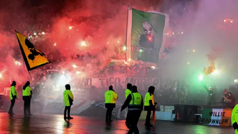 SNS Smoke and red flares visible in a stand full of football fans. Multiple stewards and police officers wearing high vis jackets are visible. Two large flags emerge from the crowd, one is yellow and black with a skull on it and the other is green and with a skeleton on it.