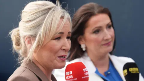 PA Media Michelle O'Neill wearing a brown blazer, she is holding a red BBC News Northern Ireland microphone, she has blonde hair.
Beside her is Emma Little-Pengelly, she has black hair and is wearing a white blazer with a blue top. She is holding a black BBC Sport NI microphone.