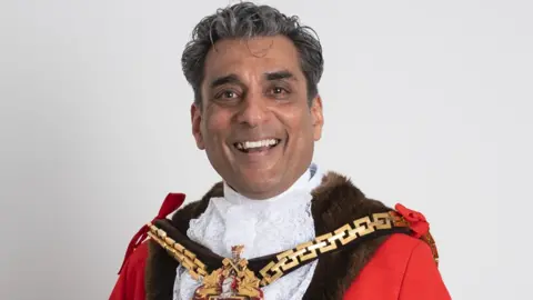 A man with dark and some grey hair is smiling at the camera. He is wearing mayoral clothing, including a mayoral chain, and standing in front of a plain white background.