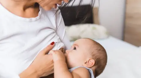 A mother breastfeeding her child. The mother is wearing a white long sleeve stop and sitting on a bed.