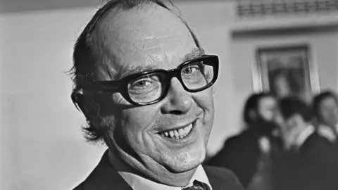 Close up of Eric Morecambe (1926 - 1984) of comedy double act Morecambe and Wise, who is wearing black glasses and smiling. The photograph is in black and white and there is a blurred image of people stood behind Eric.