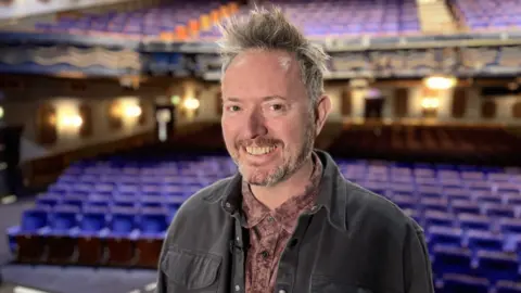 Simon Creed is pictured smiling in a theatre with purple seats. He has spiky grey hair, wears a washed purple denim shirt and a dark grey denim jacket.