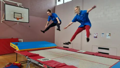 St Andrews Healthcare Two women in blue t-shirts mid air following a jump on a wide trampoline with red and blue trim. 