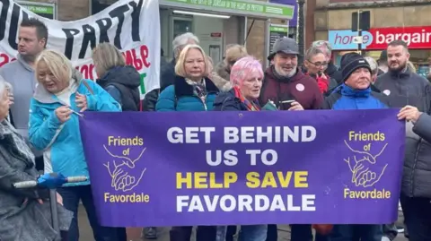 About a dozen people stand behind a purple banner saying "Get behind us to help save Favordale"