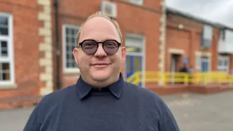 A man wearing a navy jumper and black glasses smiling, he has light blonde hair and a beard. 