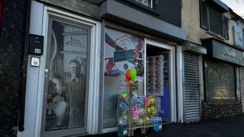 A street view of a shop front. The shop door is open and a box with football is out front. A picture of a man on a glass door can be seen to the left of the shop. A shop to the right has its shutters down.