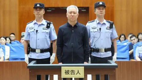 China Evergrande Group founder Hui Ka Yan takes the stand in a court in China. He has a blank look and is standing between two officers, with members of the audience in the background.