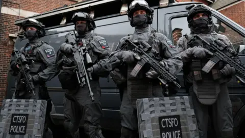 PA Media Four armed police officers in body armour, masks and helmets standing in front of a grey van.