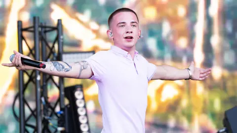 Getty Images ArrDee looking at the crowd with his arms spread out with a mic in his right hand. he's wearing a light pink tshirt with a collar. He has an ear piece in his ears. Behind him is staging with multi coloured lights and metal railing holding up light and sound equipment.