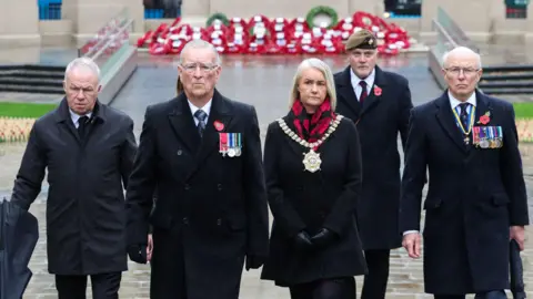 Tracy Kelly is wearing the mayoral chain of Belfast. She has shoulder length blonde hair and is dressed in black with a red and black scarf. One either side of her are two men dressed in black overcoats decorated with poppies and military medals. There are two other men also dressed in black, one of whom is wearing a military beret. In the background there are several rows of poppy wreathes.