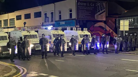 BBC A line of police officers in riot gear, with shields, stand in front of a row of armoured police Land Rovers