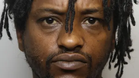 Mugshot of Ismail Hussain, standing against a grey backdrop. He has dark brown eyes, short, wispy facial hair, and long dreadlocks which cover his ears.