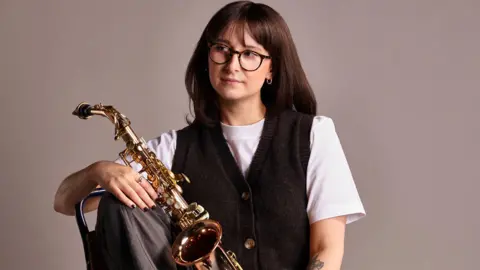 Emily Dennison A lady with brown hair and glasses is holding a saxophone looking to the left