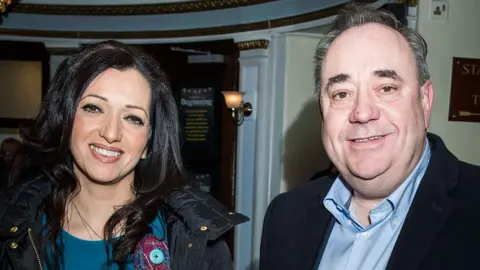 Tasmina Ahmed-Sheikh, wearing a blue top and black coat, stands beside Alex Salmond, wearing a blue shirt and dark jacket