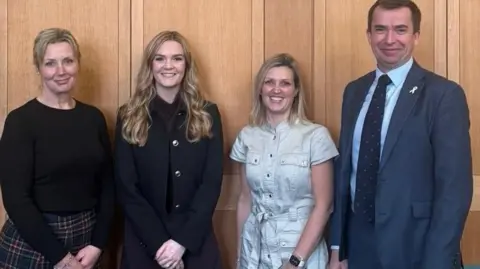 Handout Three women and a man stand against a wood panelled wall. They are smiling and looking at the camera.