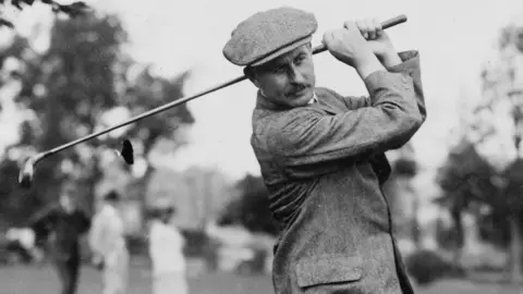Harry Vardon Trust A black and white picture of Harry Vardon with a golf club. He's wearing a flat cap and tweed jacket. 
