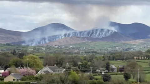 Beyond country houses, hedges, trees and rolling hills, a black mountaintop smoulders, with streaks of smoke rising into the sky. 