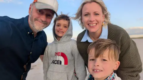 Helen Marsden A family group with a man with a baseball cap on and a beard and a women with wavy blonde hair and two boys smiling into the camera. It looks like they are on a beach somewhere.