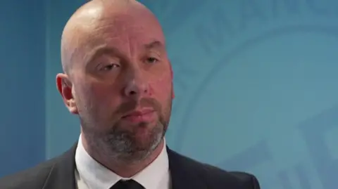 Detective Russ Clarke stands in front of a blue-coloured backdrop bearing the Greater Manchester Police logo. He is wearing a suit. 