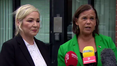 Michelle O'Neill, a woman with shoulder-length blonde hair, standing beside Mary Lou McDonald during a Sinn Féin press conference.  McDonalds has short, dark hair andis wearing a black top and a bright green blazer.  O'Neill is weaing a white blouse and black suit jacket. 