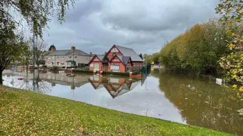 Lea Brook church A red brick church and a few houses sit surrounded by water. Their reflections can also bee seen in the water. The water stretches across a road up to a grass verge covered in leaves. Leaves are also floating in the water. The church is on a corner with another water covered road next to it. An emergency vehicle and a figure can be seen in the distance