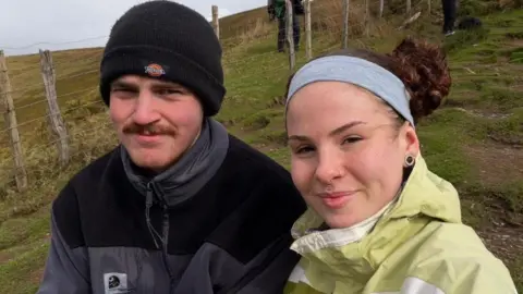 Supplied Justyce and David hiking in a moorland. Both are wearing coats. Justyce has her hair in a grey headband. David is wearing a woolly hat.