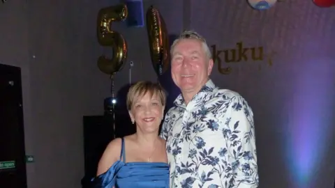 Family photo Marianne and her husband at her 50th birthday party. Marianne is wearing a blue strappy dress and her husband is wearing a white short with a blue floral print. There are two balloons - a five and a zero - behind them. 