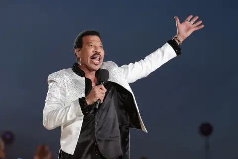 Reuters Lionel Richie in a white, jewel-encrusted jacket, raises a hand to the air as he performs in concert
